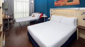 Habitación de hotel con una cama grande, una cama más pequeña, un escritorio con silla, una mesita de noche, una lámpara y un cuadro en la pared.