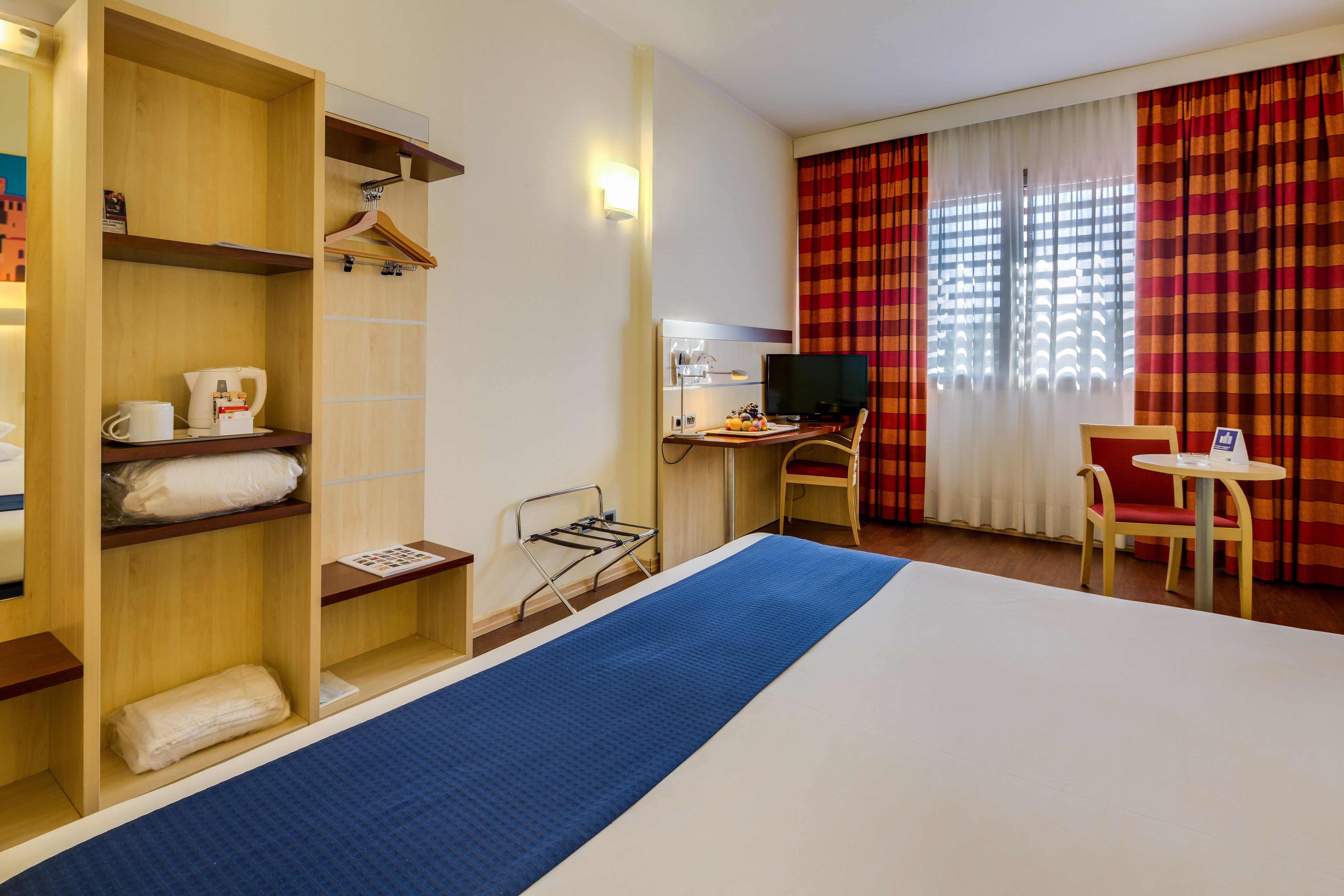 A hotel room with a bed, a desk with a computer, a chair, a small table, a TV, and a wardrobe with shelves.
