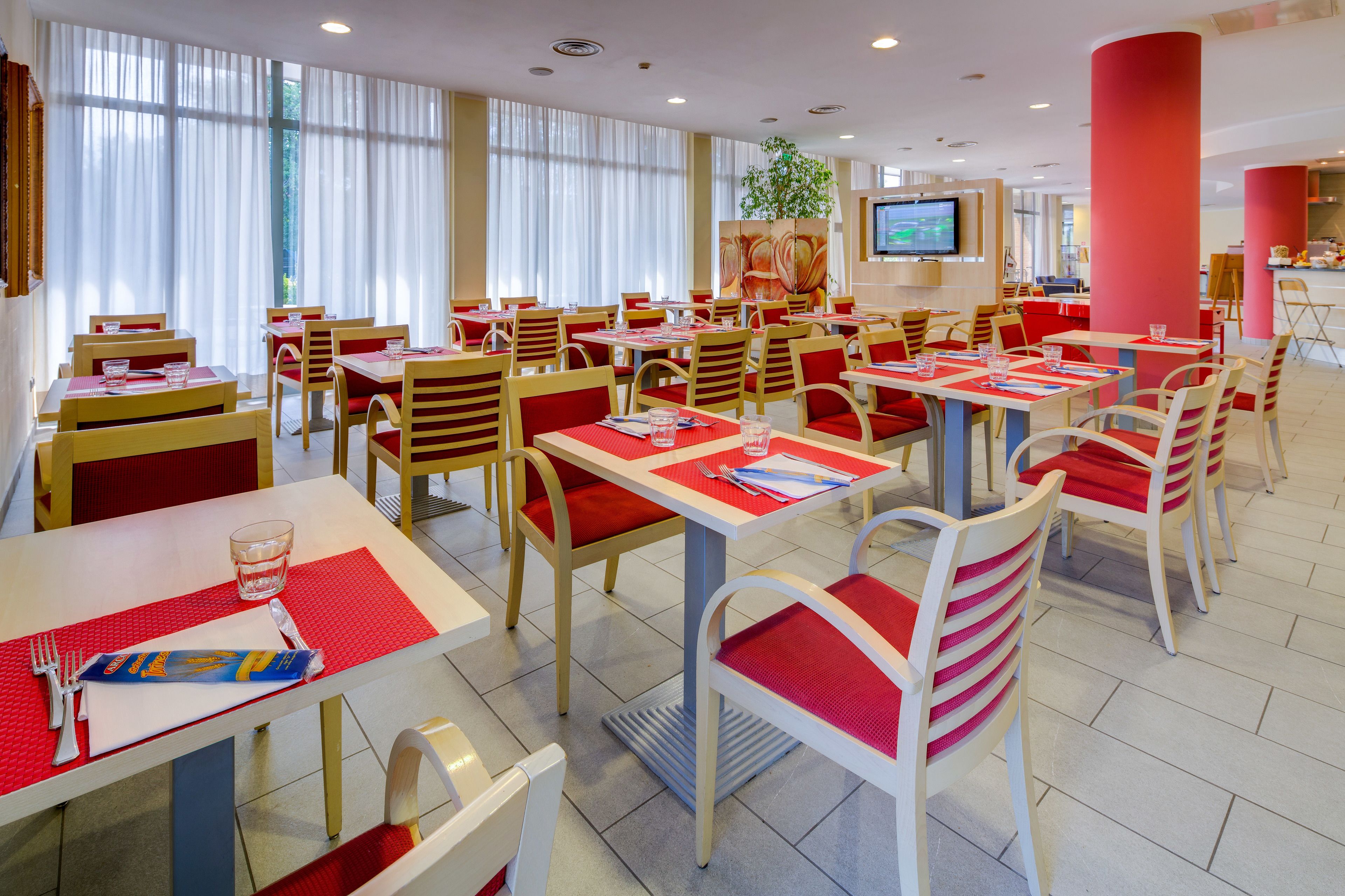 A spacious restaurant with multiple tables and chairs, a television screen, and a large window with curtains.