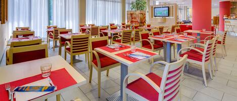 A spacious restaurant with multiple tables and chairs, a television screen, and a large window with curtains.