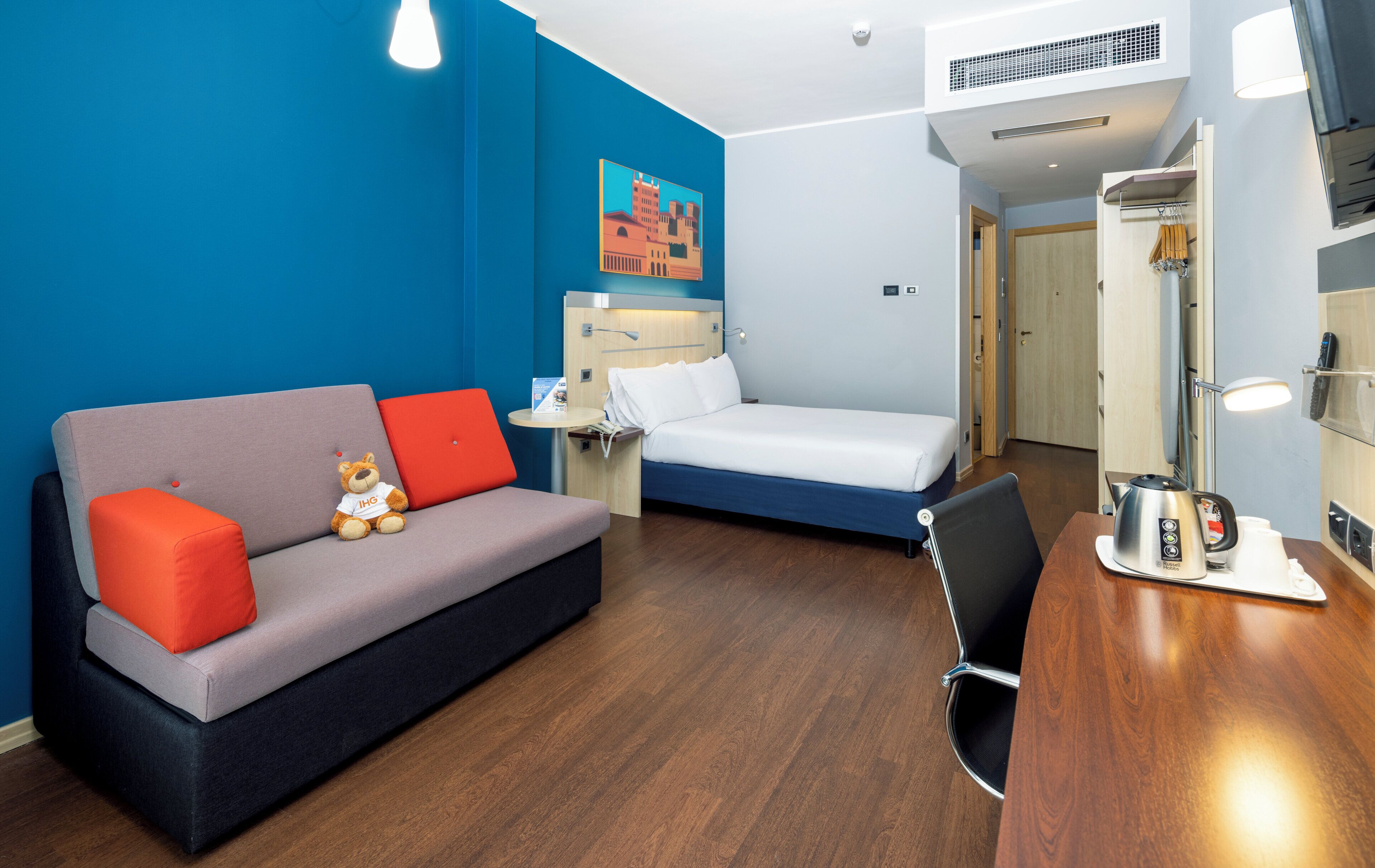A compact hotel room with a grey sofa, a single bed, a wooden desk, a small table, and a wall-mounted air conditioner.
