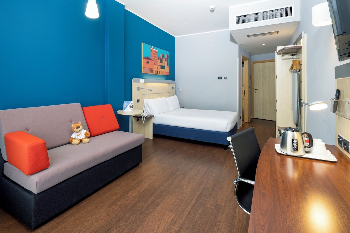 A compact hotel room with a grey sofa, a single bed, a wooden desk, a small table, and a wall-mounted air conditioner.