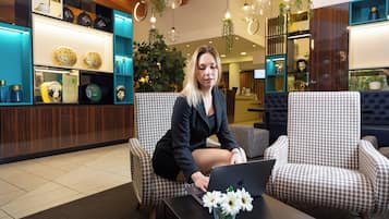 Una mujer sentada en una silla estampada en un vestíbulo, con una computadora portátil, un jarrón de flores y una exhibición de objetos decorativos en estanterías.