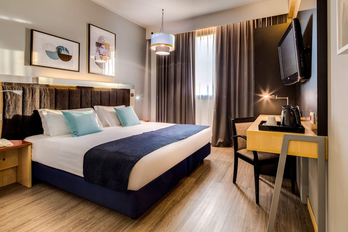 A hotel room with a large bed, a desk, a chair, a television, and artwork on the wall.