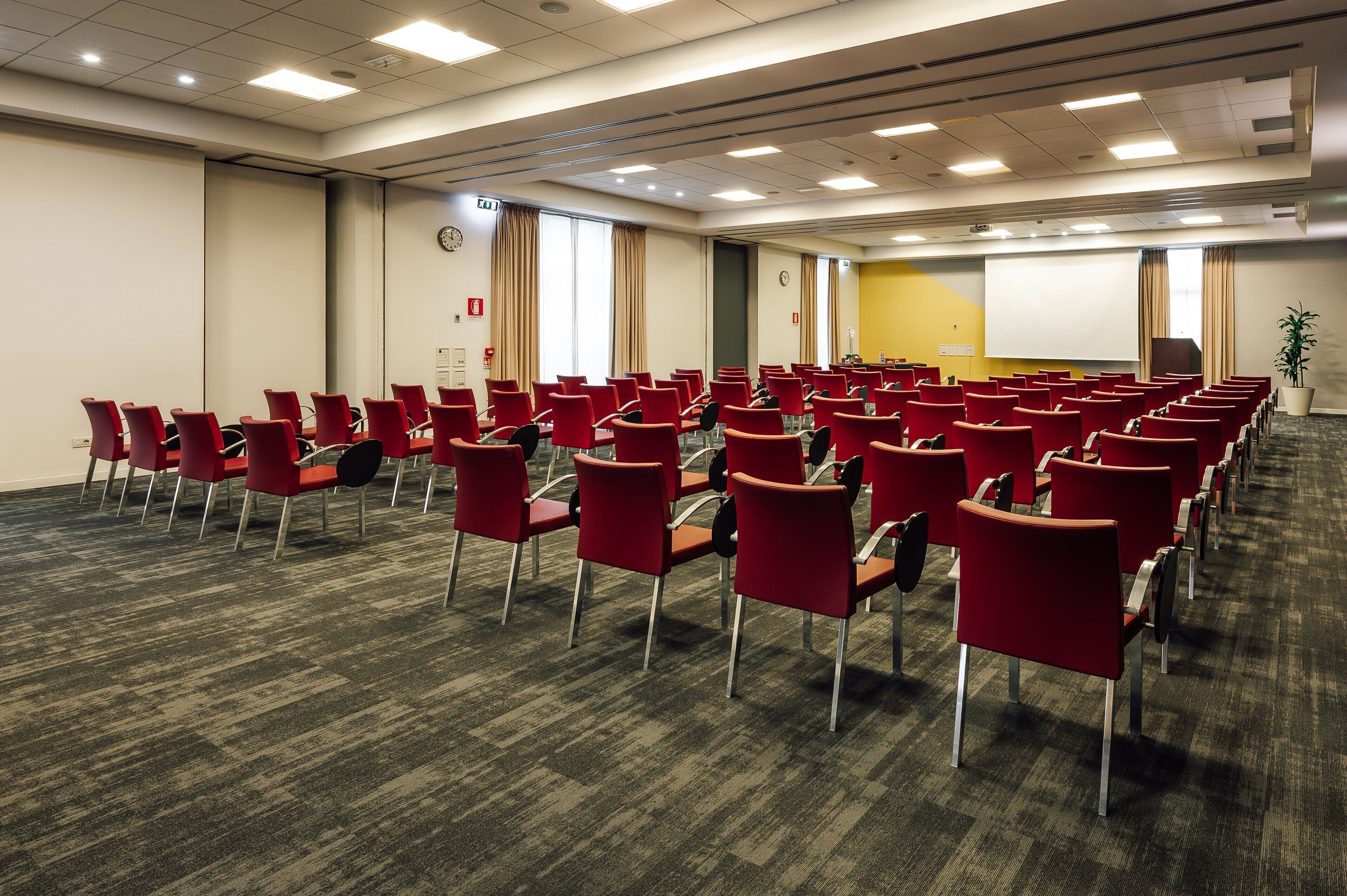 Ein Konferenzraum mit Reihen roter Stühle, einer Projektionsleinwand und einem Teppichboden.