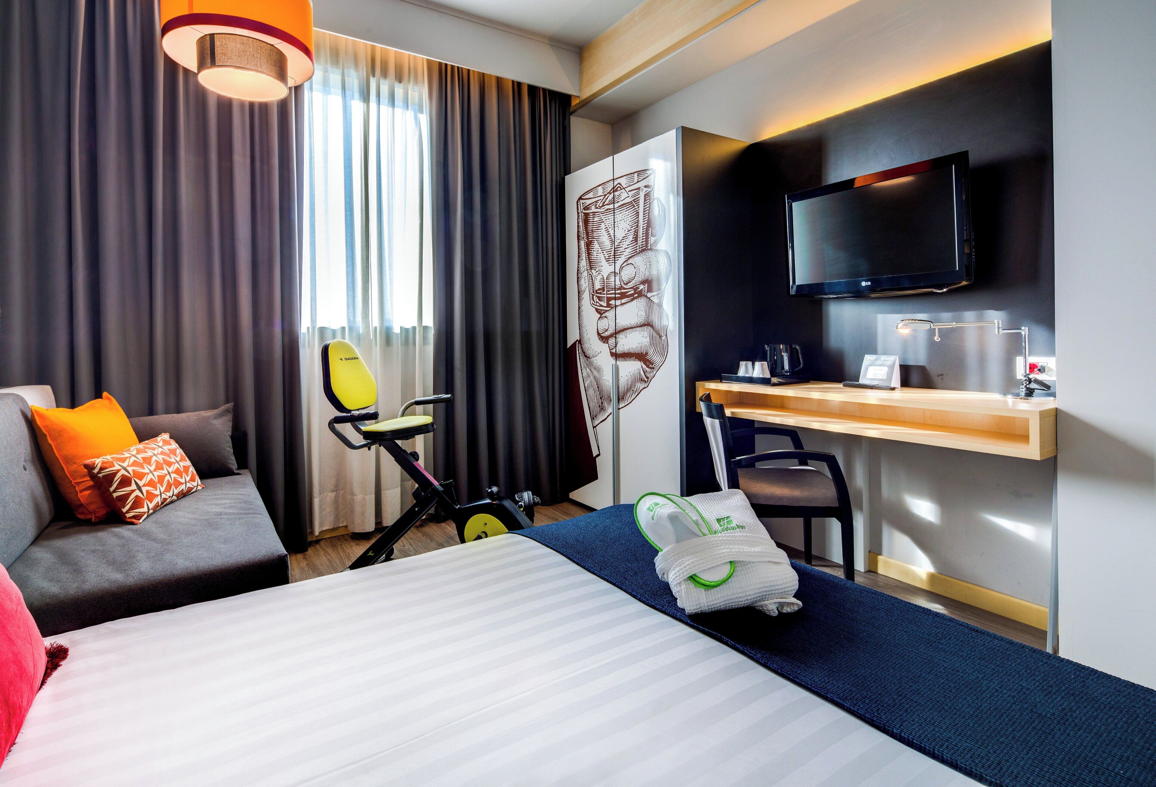 A hotel room with a bed, a desk with a flat-screen TV, a chair, a treadmill, and a window with curtains.