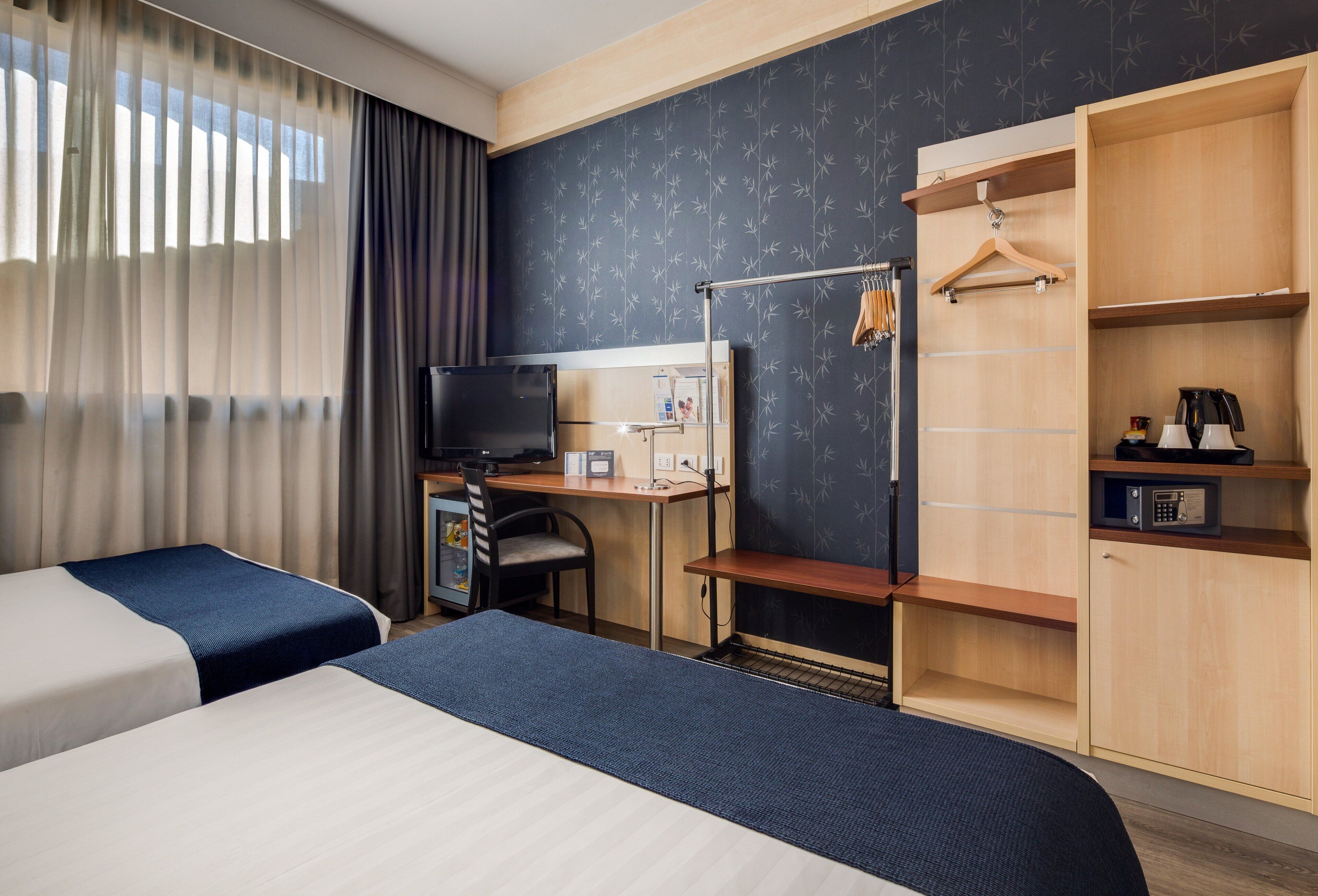 Habitación de hotel con dos camas, un escritorio con computadora, un televisor y un armario con perchero.