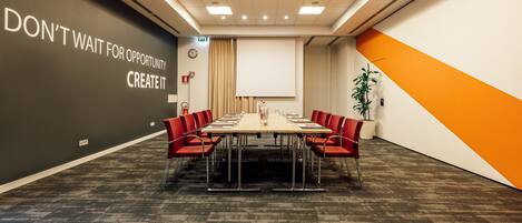 A conference room with a long table, red chairs, a projector screen, and motivational wall art.