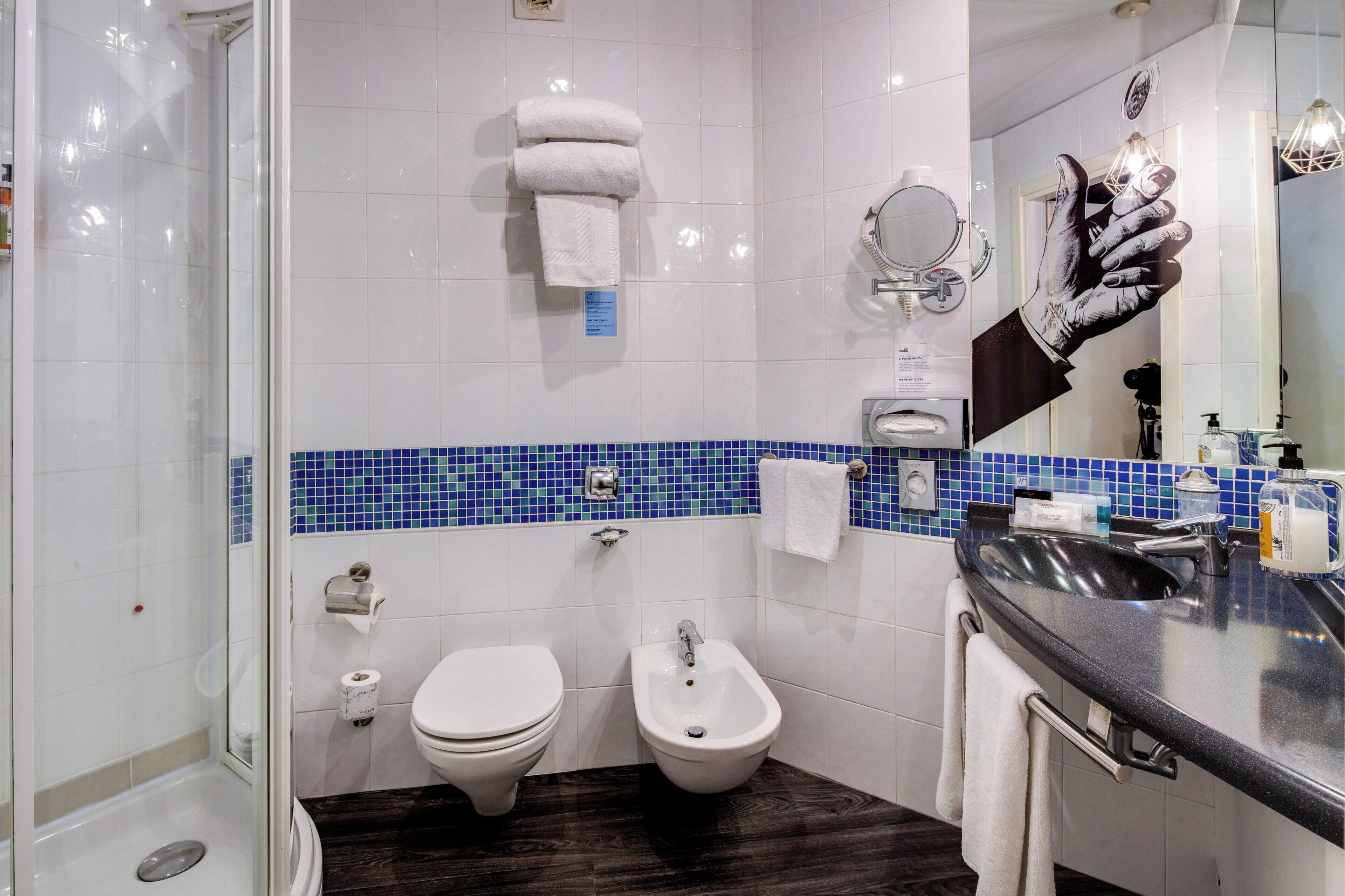 Un baño moderno con inodoro, bidé y lavamanos con espejo. Hay toallas colgadas en un perchero y un dispensador de gel antibacterial en el lavamanos.
