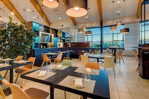 A modern restaurant with high ceilings, exposed beams, and large windows. Tables are set with placemats, cutlery, and glasses. - Holiday Inn Bologna - Fiera by IHG (Bologna)