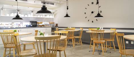 Un restaurante con mesas redondas y sillas de madera, una cocina al fondo y un reloj de pared.