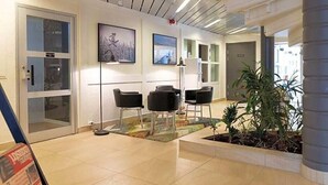 Un área de recepción con puerta de vidrio, sillas negras, una lámpara de pie y cuadros en la pared.