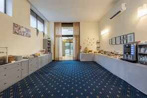 Une chambre d’hôtel avec un tapis à motifs bleus, un coin petit-déjeuner équipé de machines à thé et à café, et une vue sur l’extérieur grâce à une grande fenêtre.