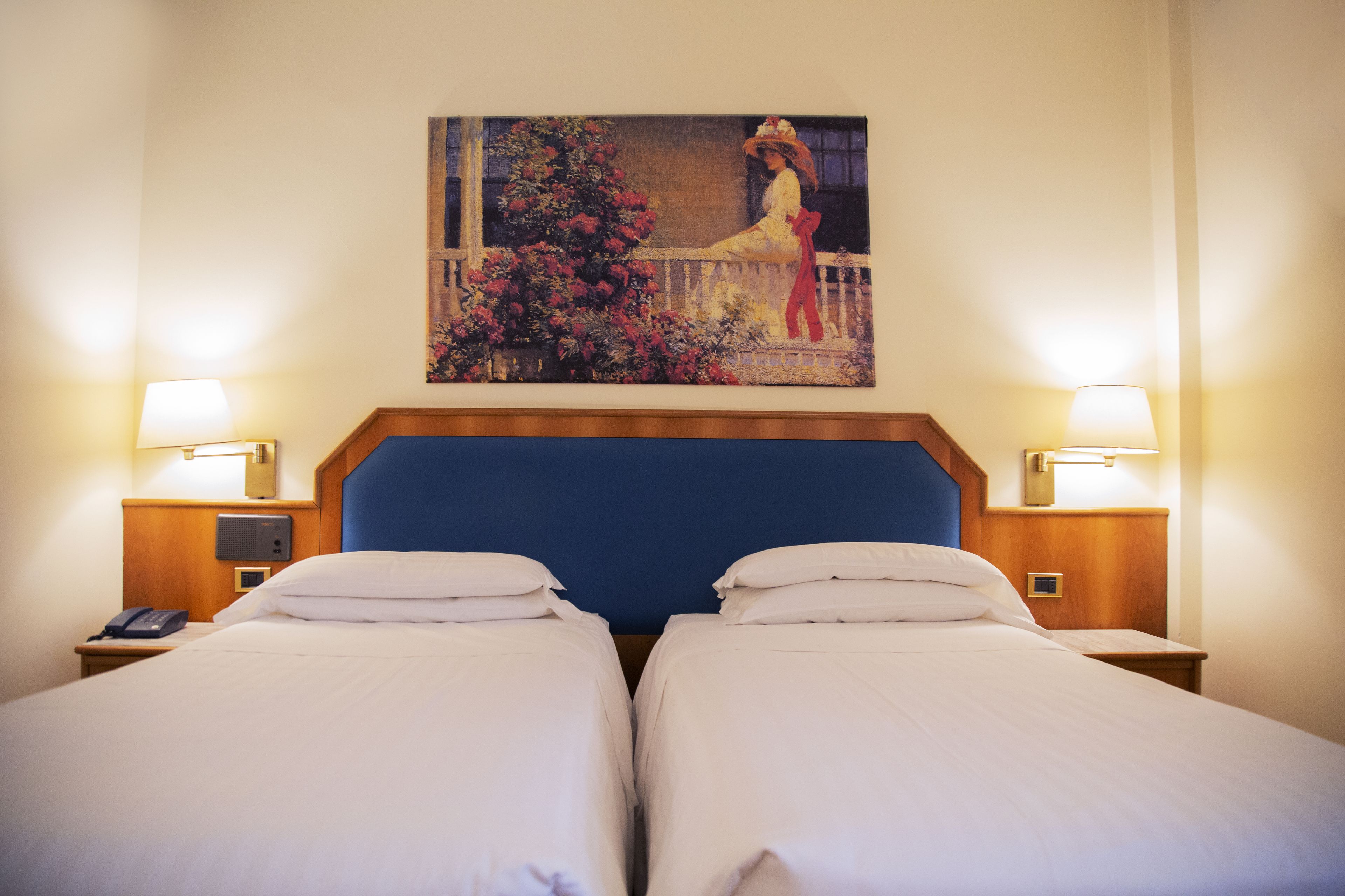 A hotel room with two beds, a painting on the wall, and bedside lamps.