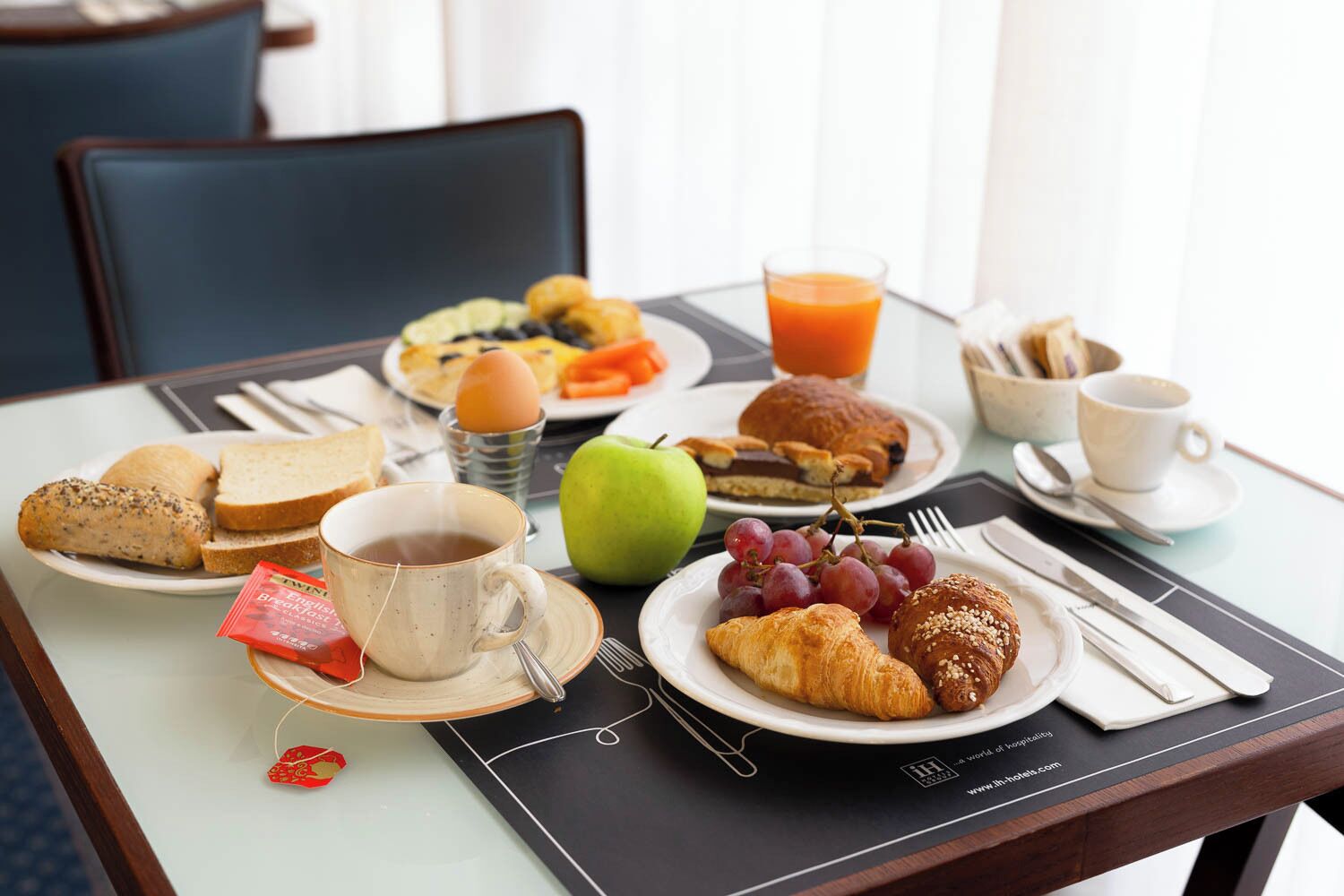 Une table de petit-déjeuner avec des croissants, du pain, une pomme, des raisins, un verre de jus d’orange, une tasse de café et une boîte de thé.