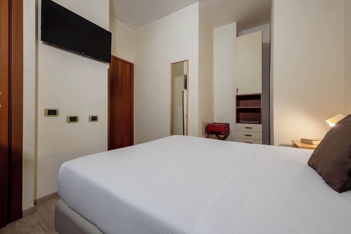 A hotel room with a bed, a wardrobe, a mirror, a dresser, and a red suitcase.