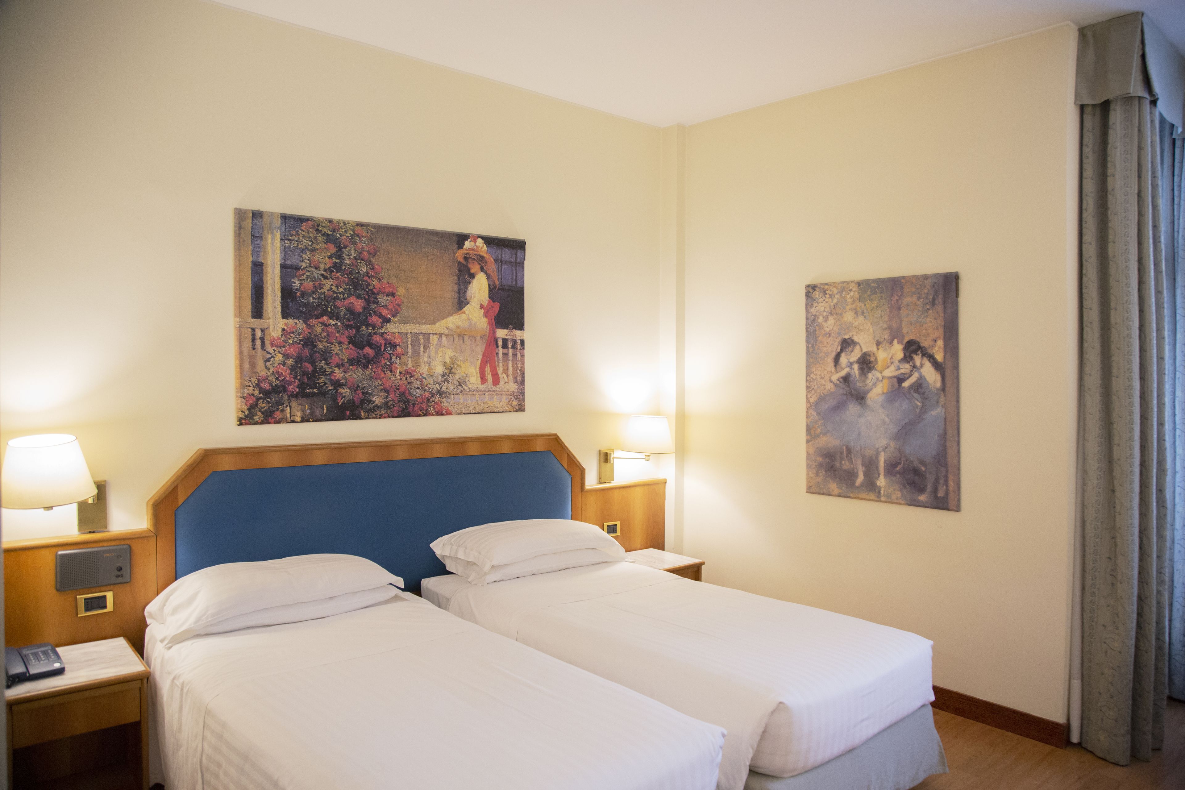 Une chambre d’hôtel avec deux lits simples, un grand tableau au mur, une table de chevet avec une lampe et une fenêtre avec des rideaux.