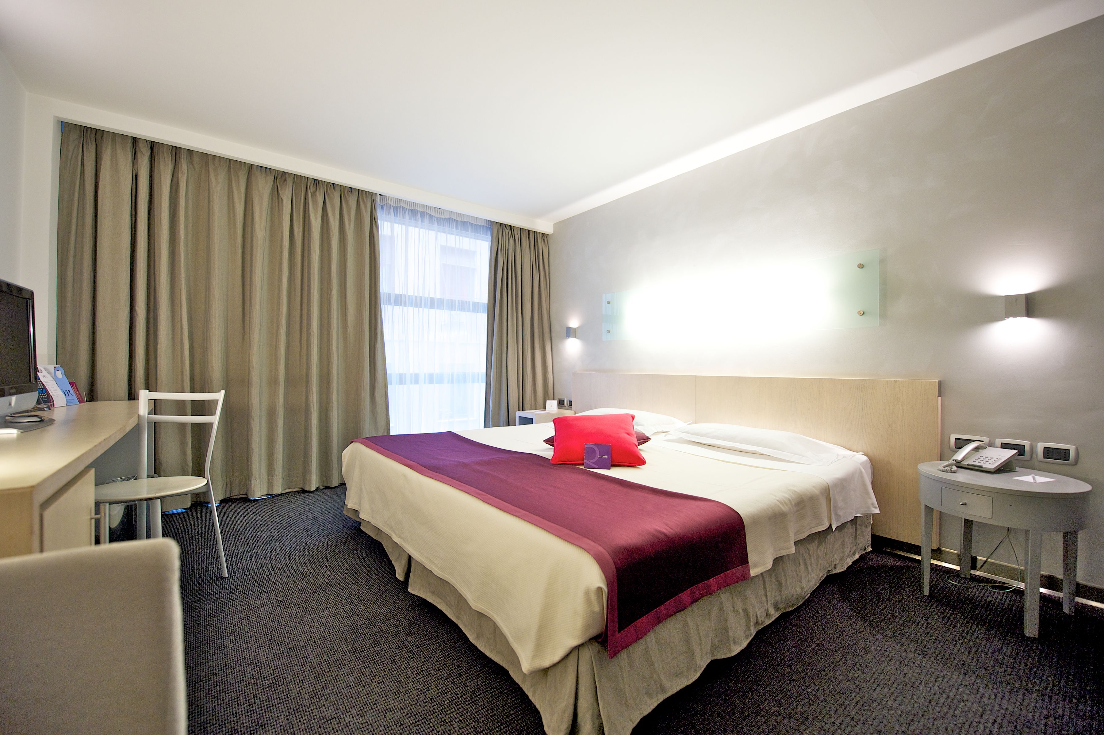A hotel room with a bed, a desk, a chair, a telephone, and a window with curtains.