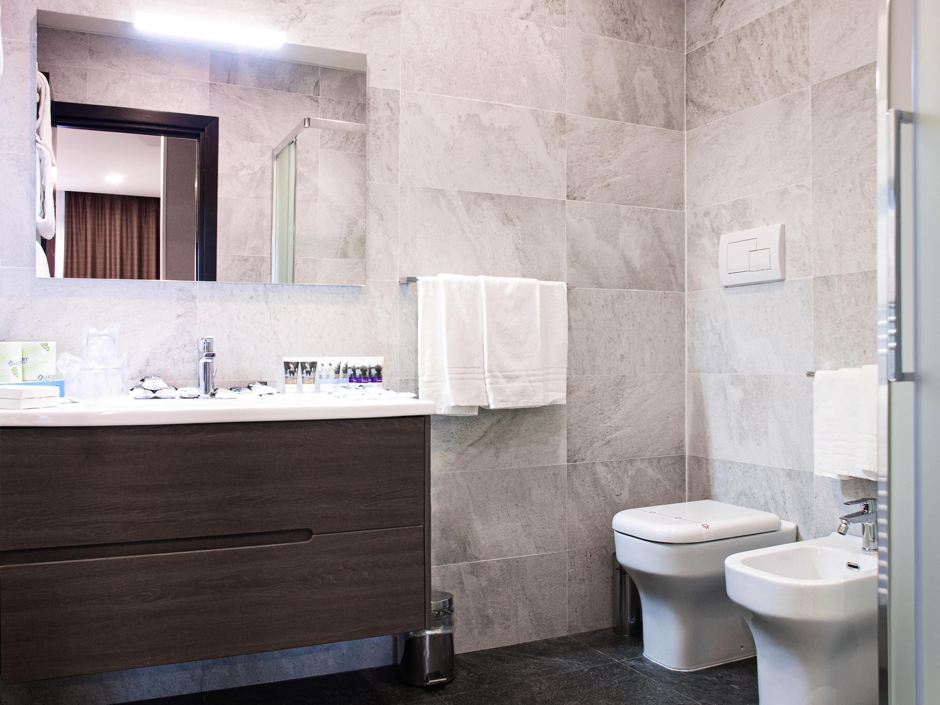 Un baño moderno con pared de azulejos de mármol, tocador de madera oscura, lavamanos blanco, espejo, inodoro y bidé.