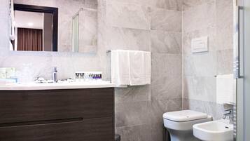 Un baño moderno con pared de azulejos de mármol, tocador de madera oscura, lavamanos blanco, espejo, inodoro y bidé.