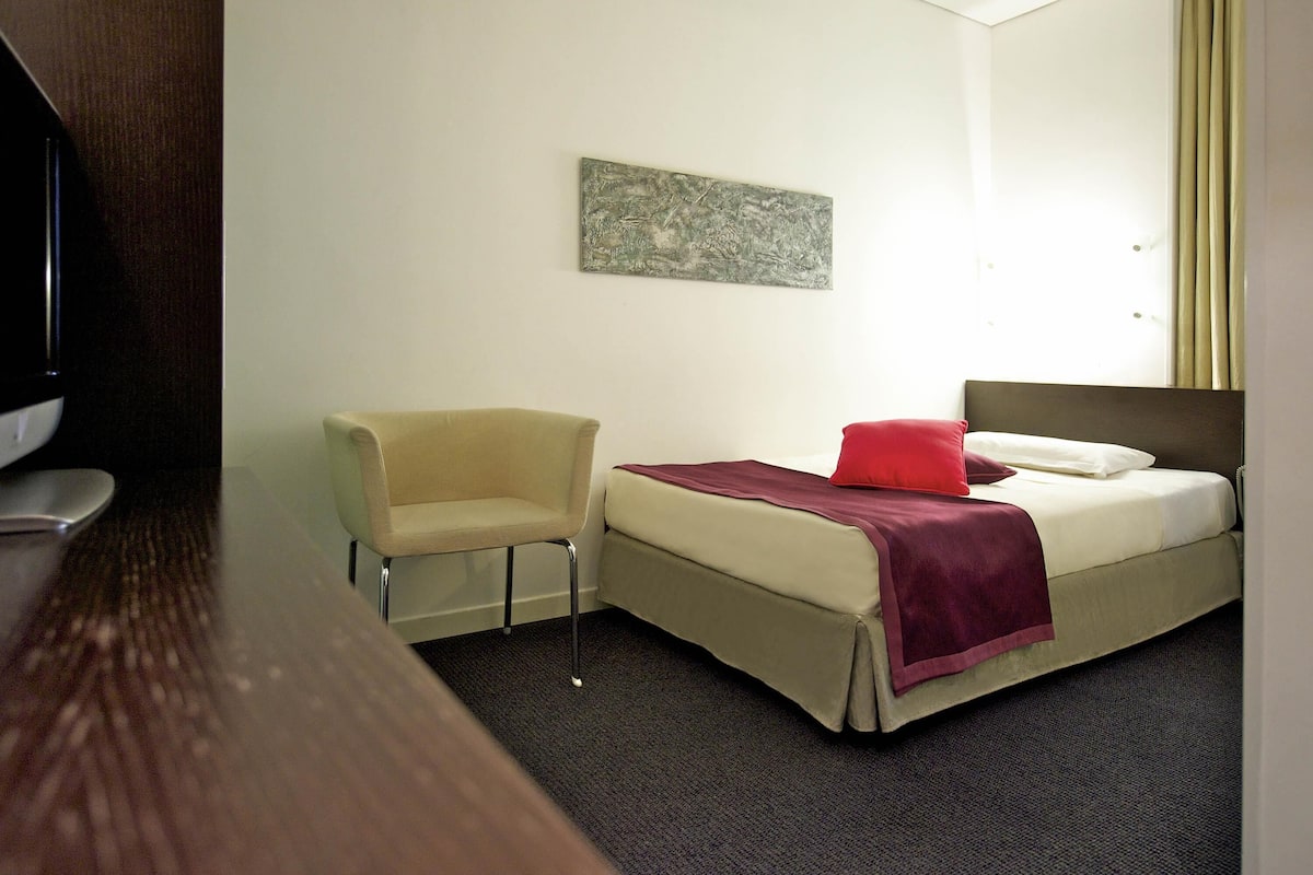 A hotel room with a bed, a chair, a television, and a painting on the wall.