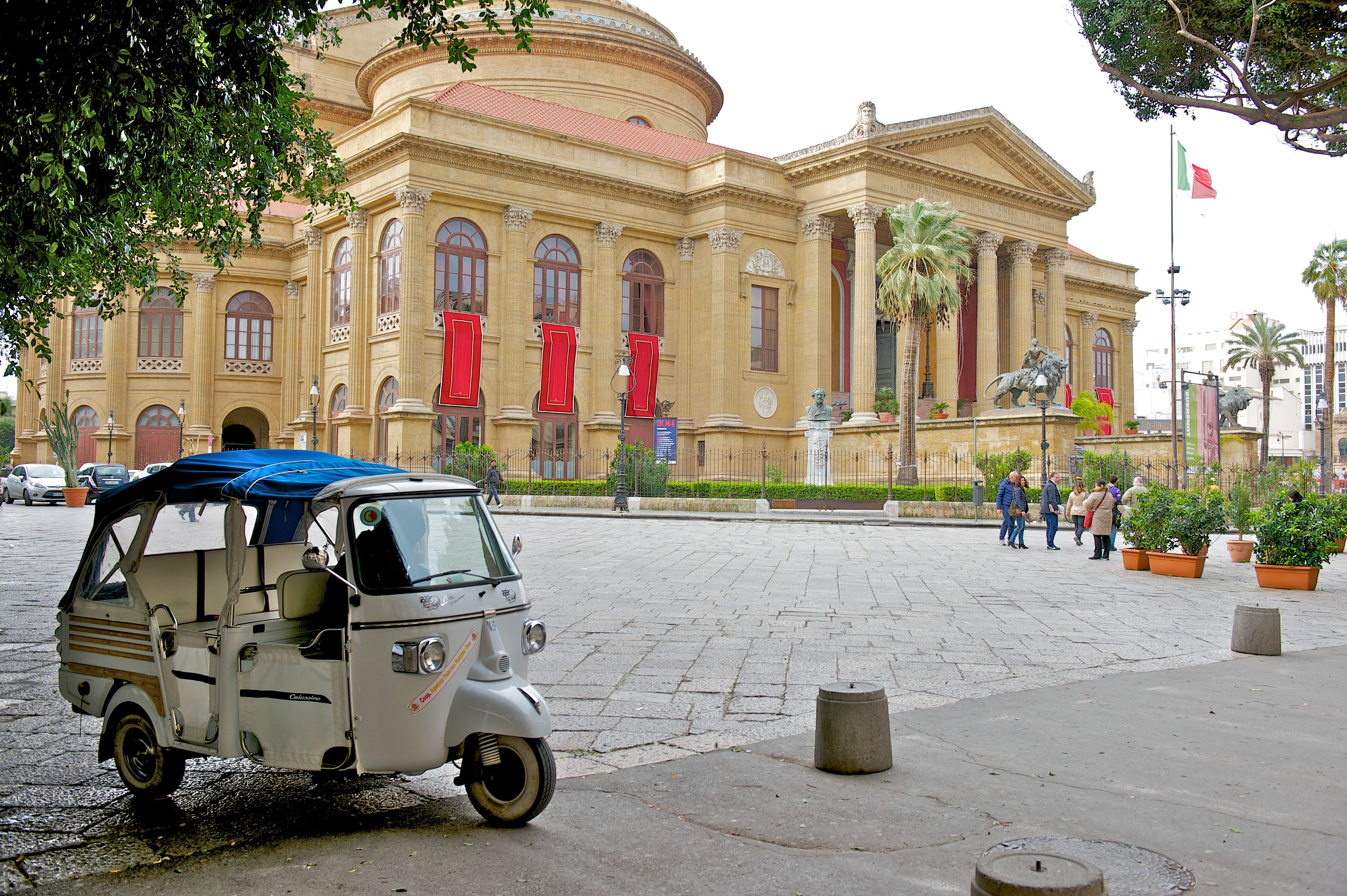 En ældre tuk-tuk parkeret foran en historisk bygning med klassisk arkitektur, palmer og et flag.