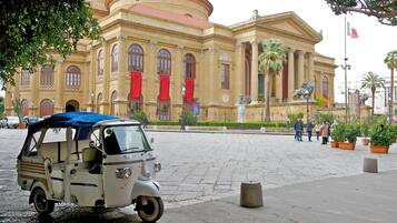 A vintage tuk-tuk parked in front of a historic building with classical architecture, palm trees, and a flag.