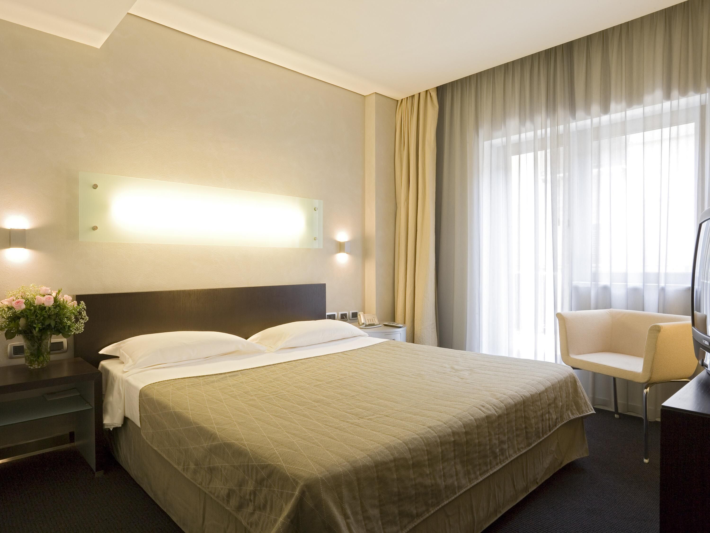 A hotel room with a large bed, a bedside table with a lamp, a chair, and a television.