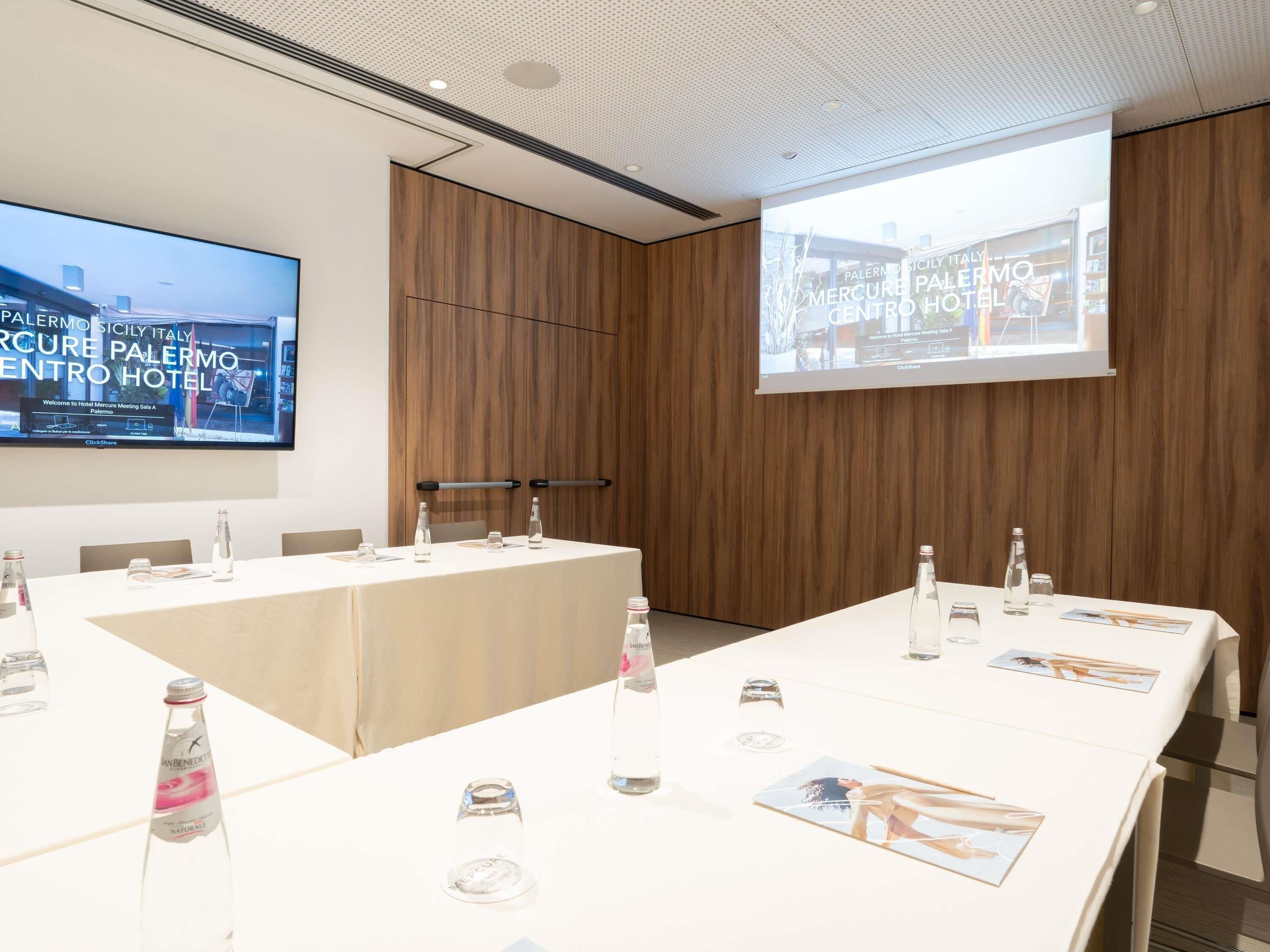 Une salle de conférence dotée d’un grand écran affichant des informations sur l’hôtel, d’une longue table avec des bouteilles d’eau et des verres, et d’un mur recouvert de panneaux en bois.