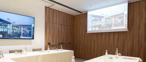 Une salle de conférence dotée d’un grand écran affichant des informations sur l’hôtel, d’une longue table avec des bouteilles d’eau et des verres, et d’un mur recouvert de panneaux en bois.