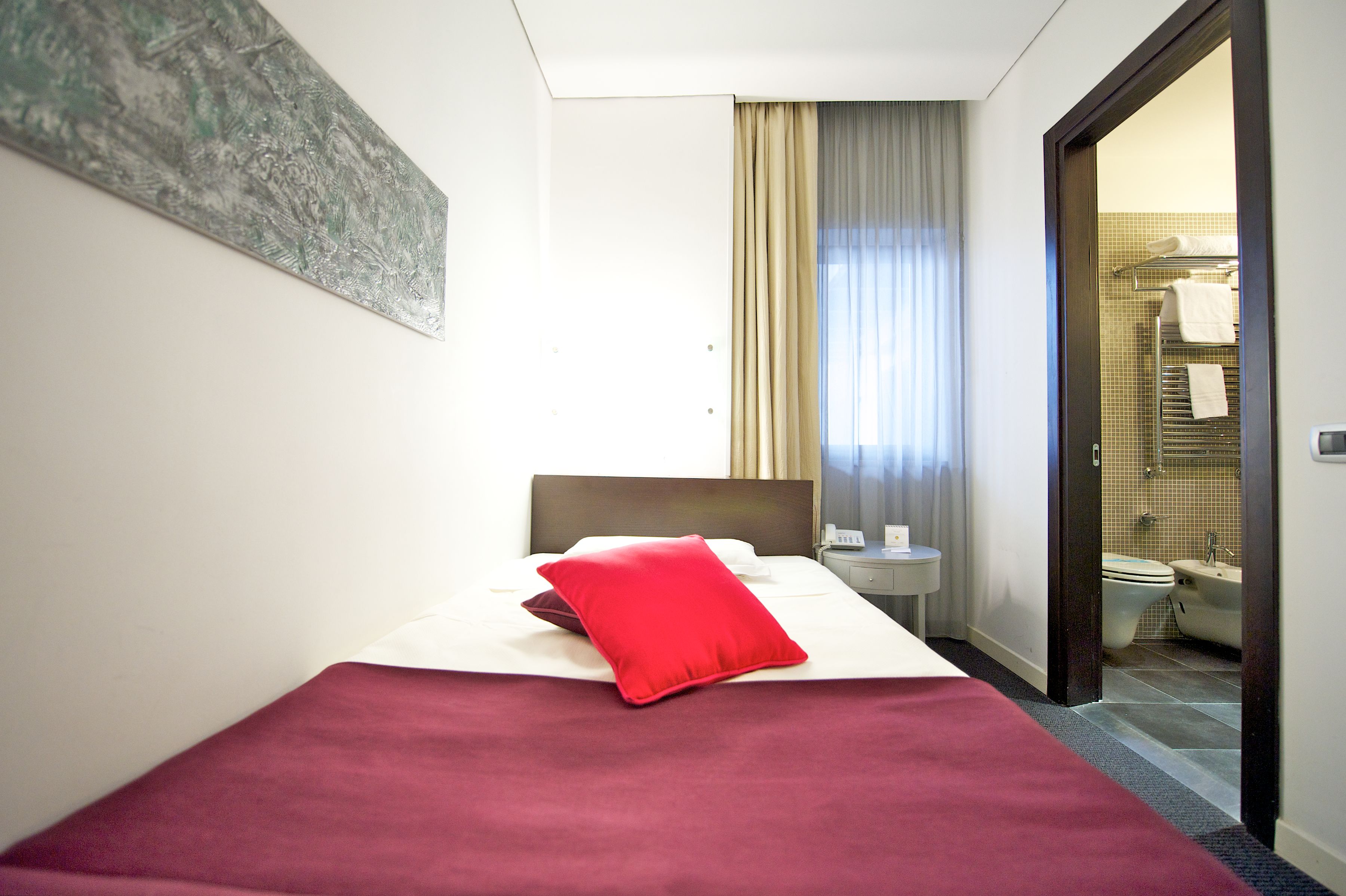 A hotel room with a bed, a red pillow, a nightstand, and a view of the bathroom.