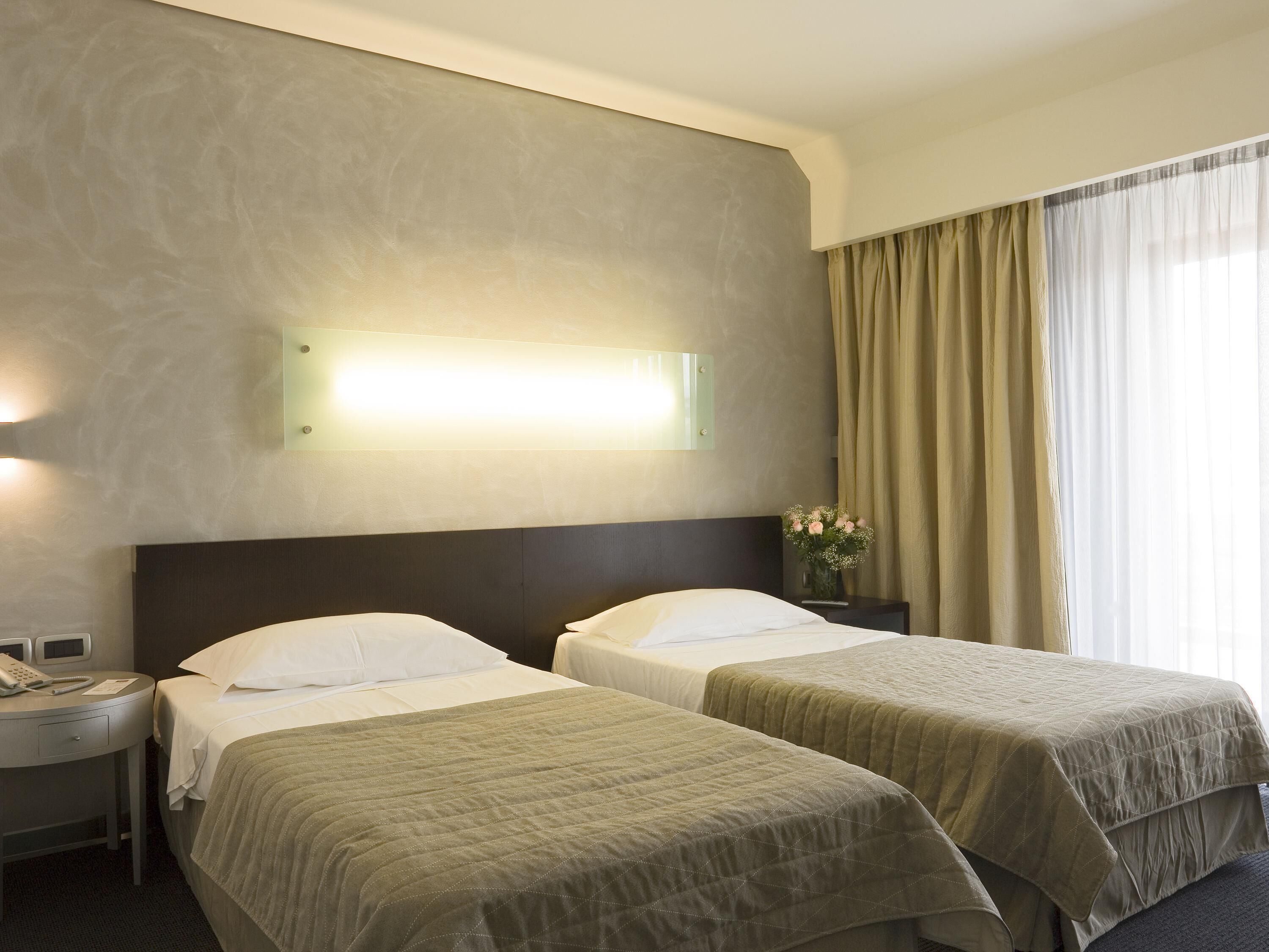 A hotel room with two beds, a bedside table with a phone, a wall-mounted light, and a window with curtains.