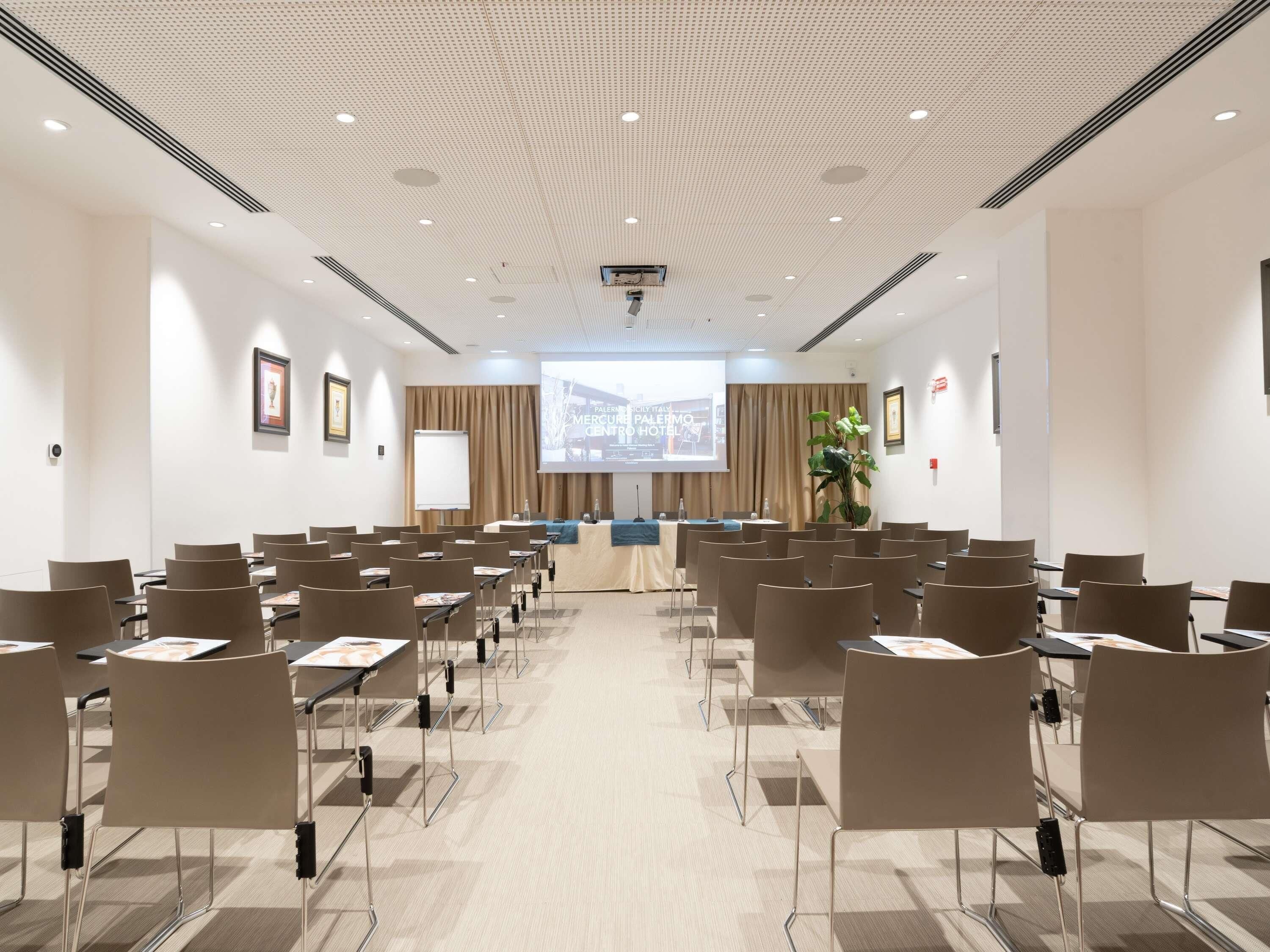 A conference room with rows of chairs, a podium, a screen, and a large plant.