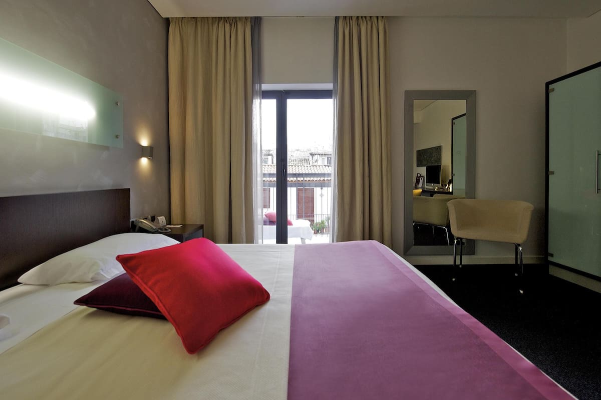 A hotel room with a bed, a desk, a chair, a window with curtains, and a door.