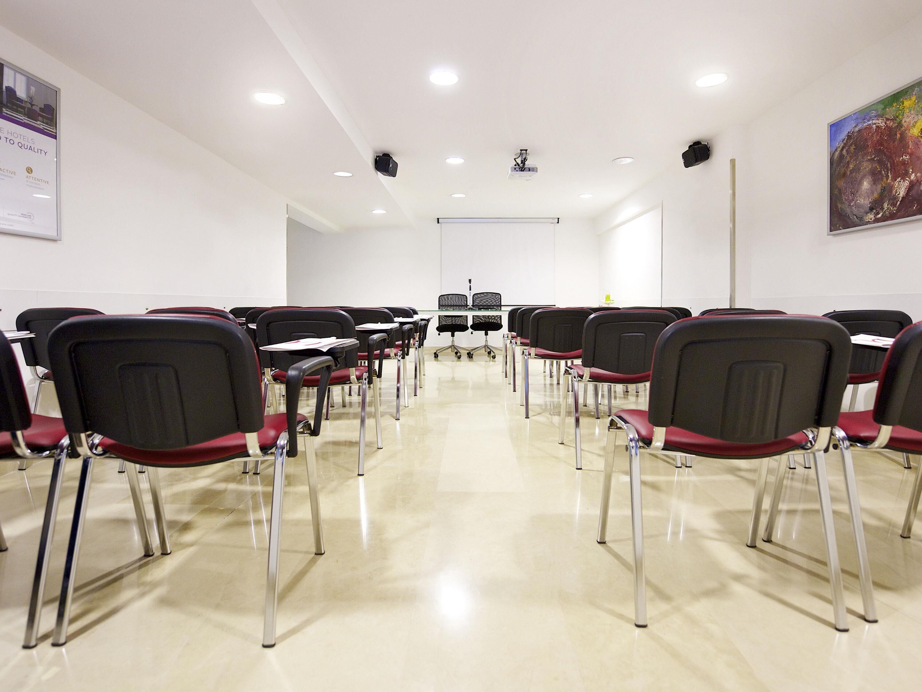 Un salón de conferencias con filas de sillas, un proyector y una pizarra.