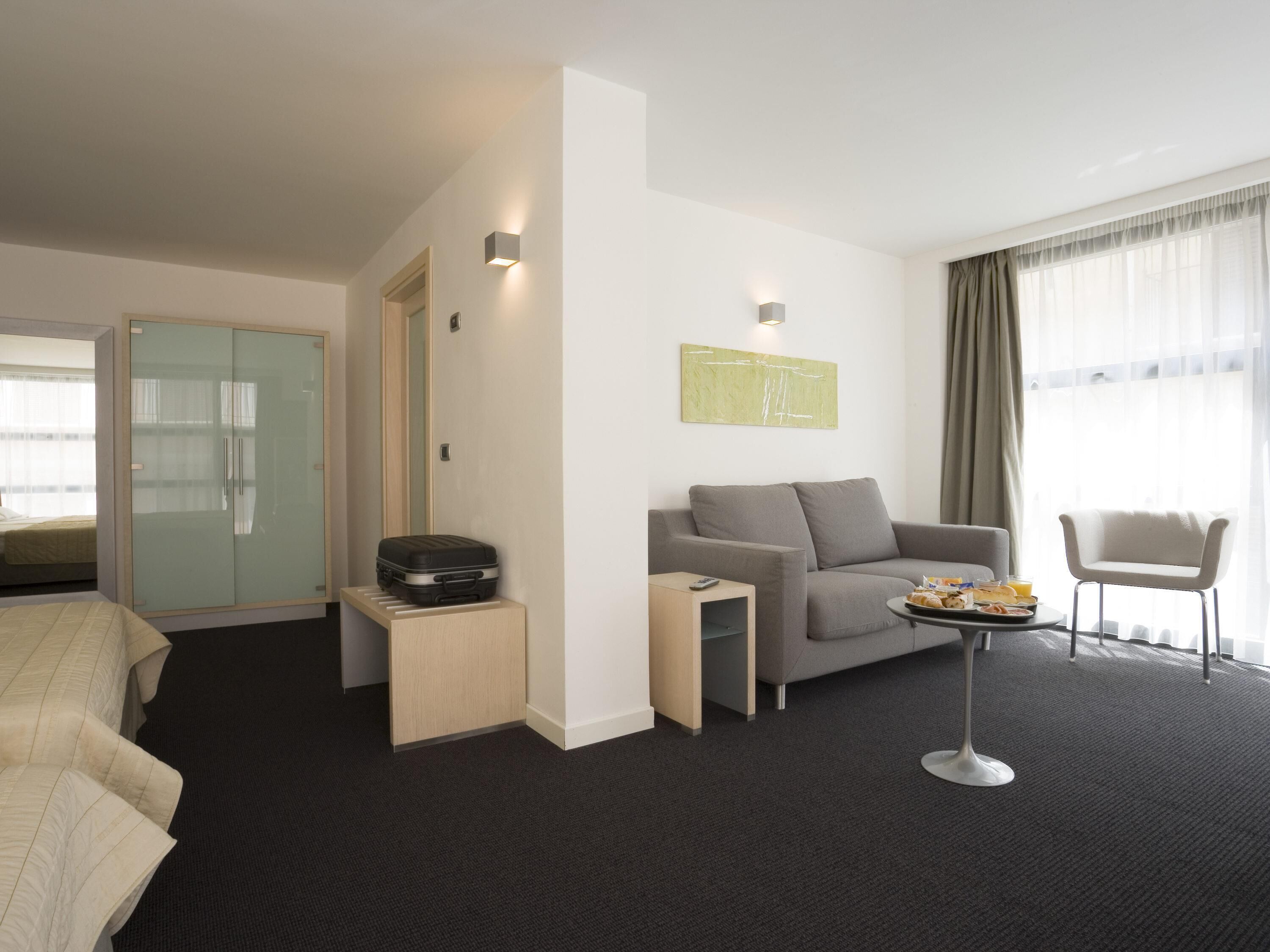 Una habitación de hotel moderna con una cama, un sofá gris, una pequeña mesa con desayuno, un armario con puerta de vidrio y un amplio ventanal con cortinas.