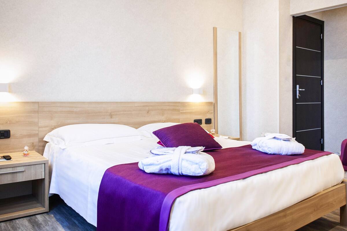 A hotel room with a wooden headboard, a bed with white bedding and a purple throw, a nightstand with a phone and a bottle, and a door with a handle.