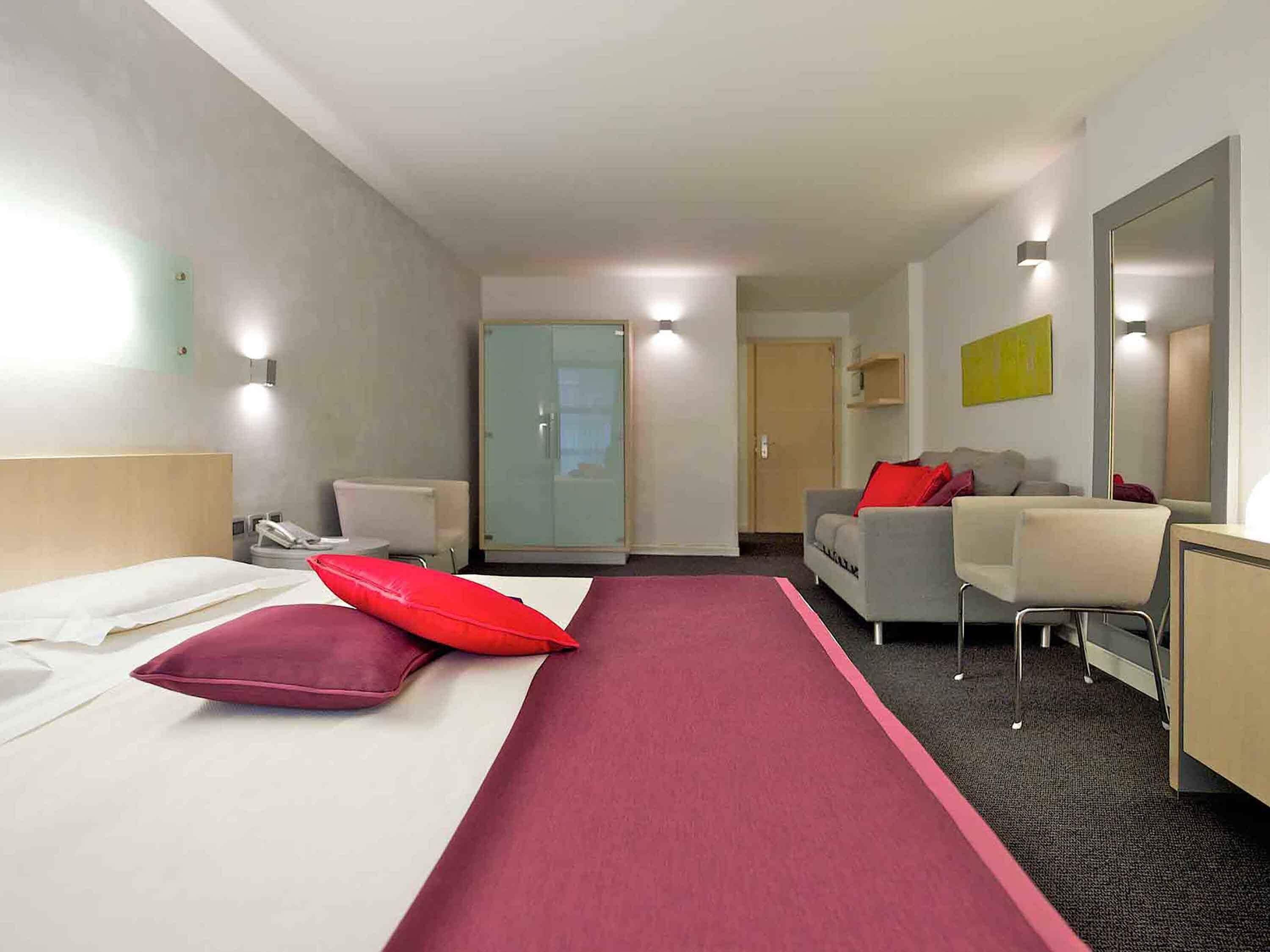 A hotel room with a bed, a desk, a chair, a sofa, a wardrobe, and a door.
