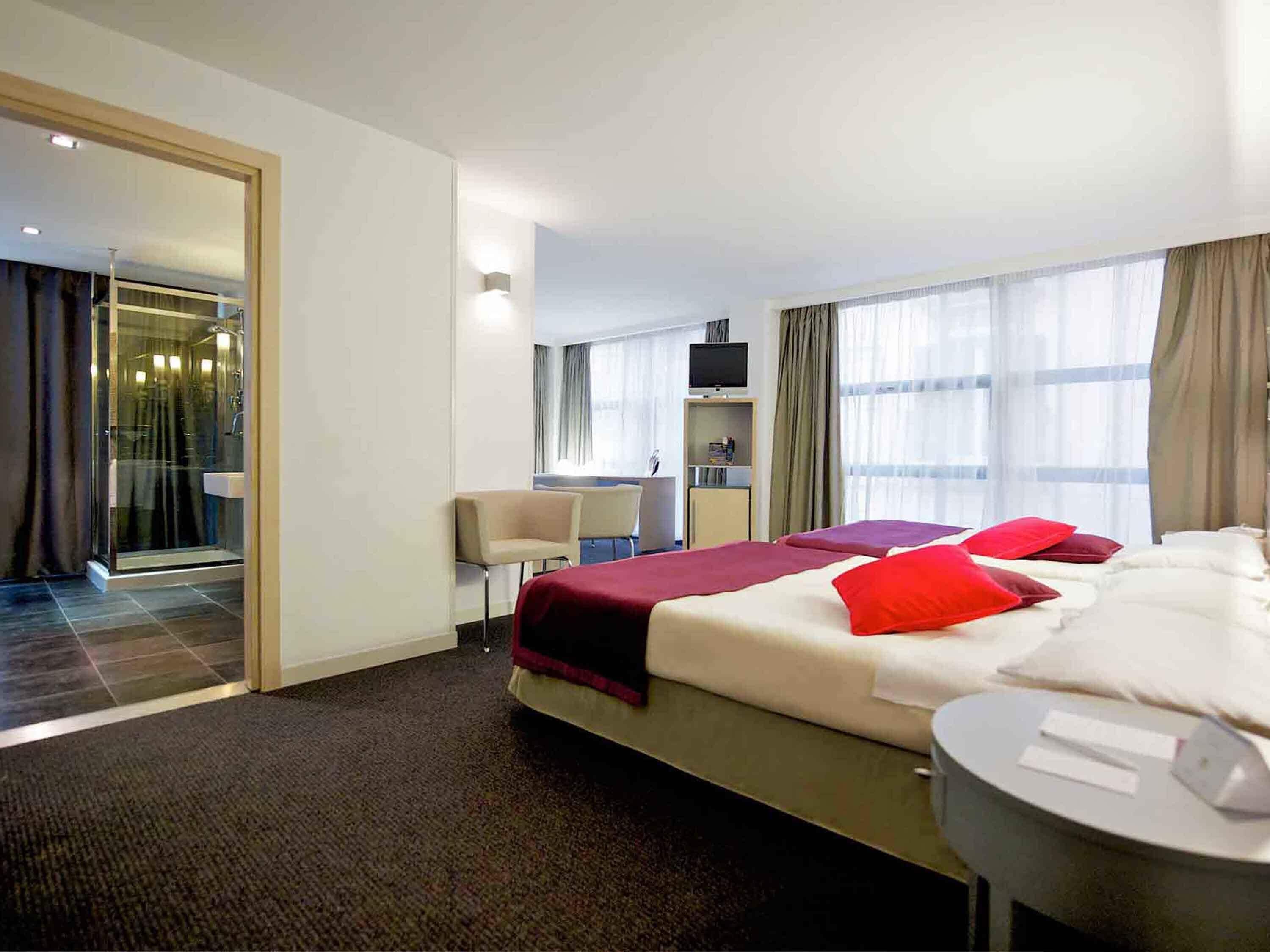 A hotel room with a bed, a desk, a TV, and a bathroom with a glass door.