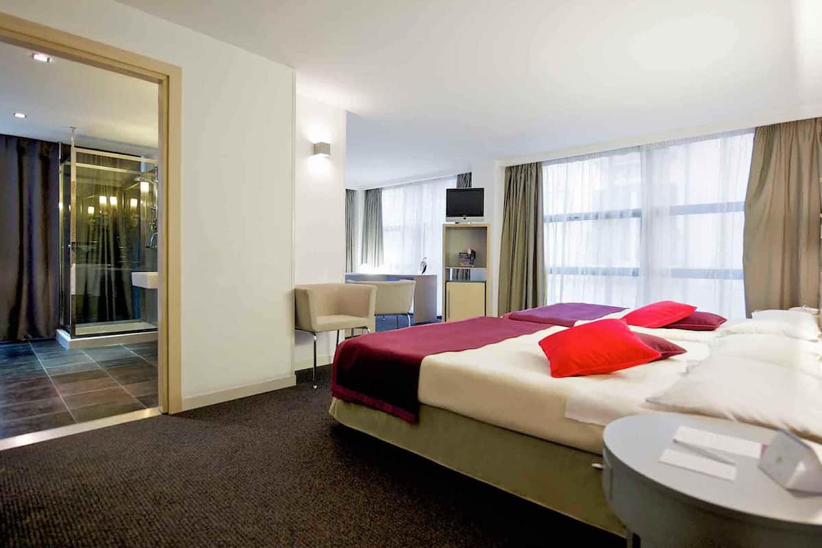 A hotel room with a bed, a desk, a TV, and a bathroom with a glass door.