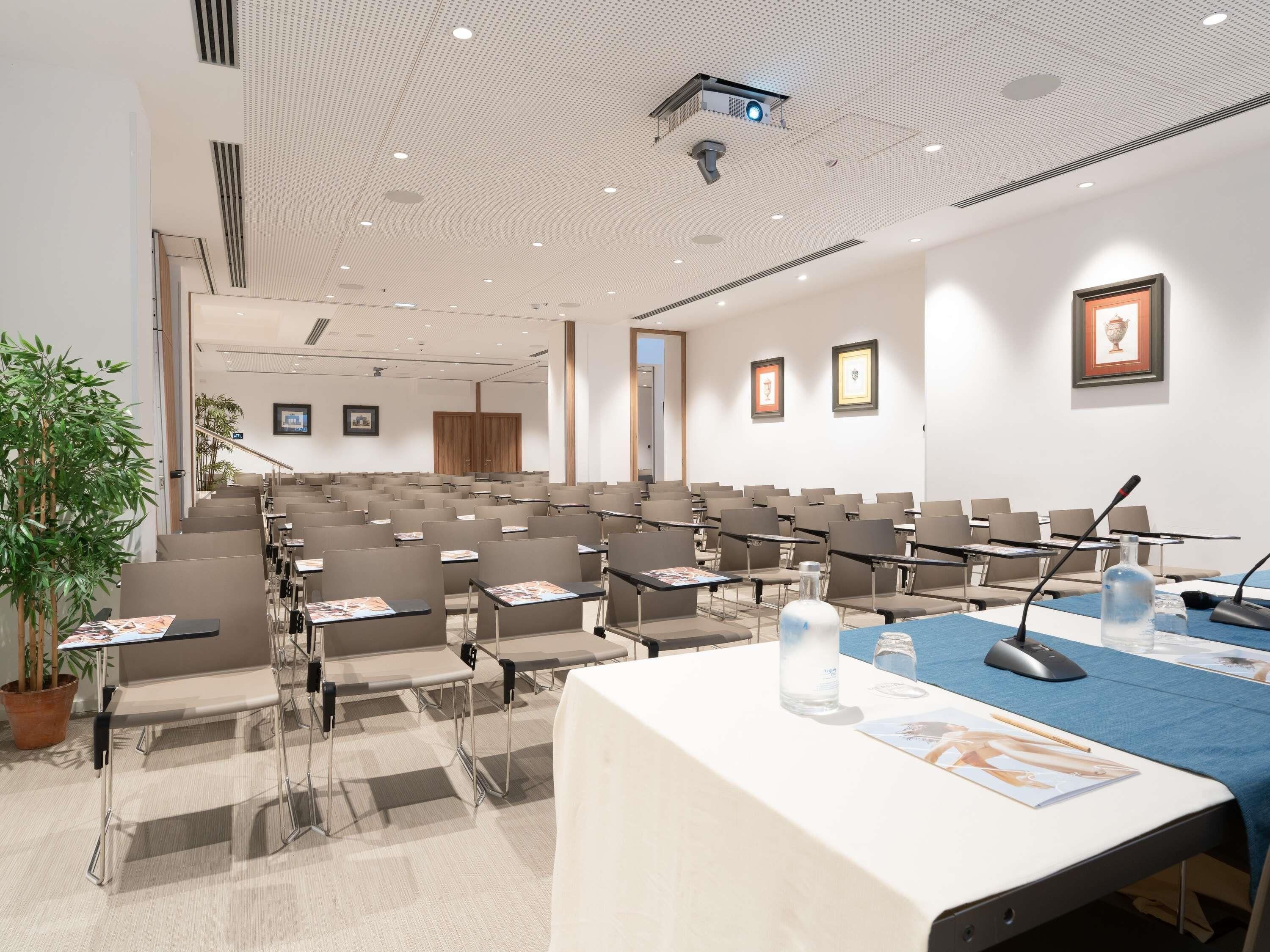 A conference room with rows of chairs, a projector, and a table with a microphone and water bottle.
