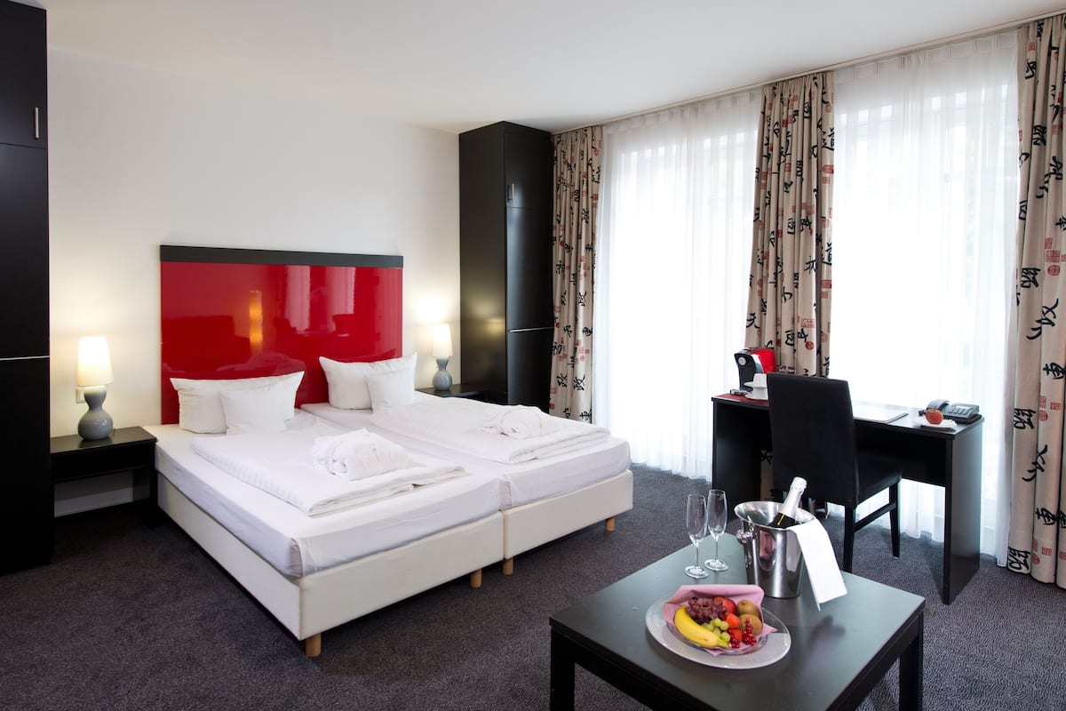 Una habitación de hotel con una cama, un escritorio, una silla, una mesa con fruta y una botella, y cortinas.