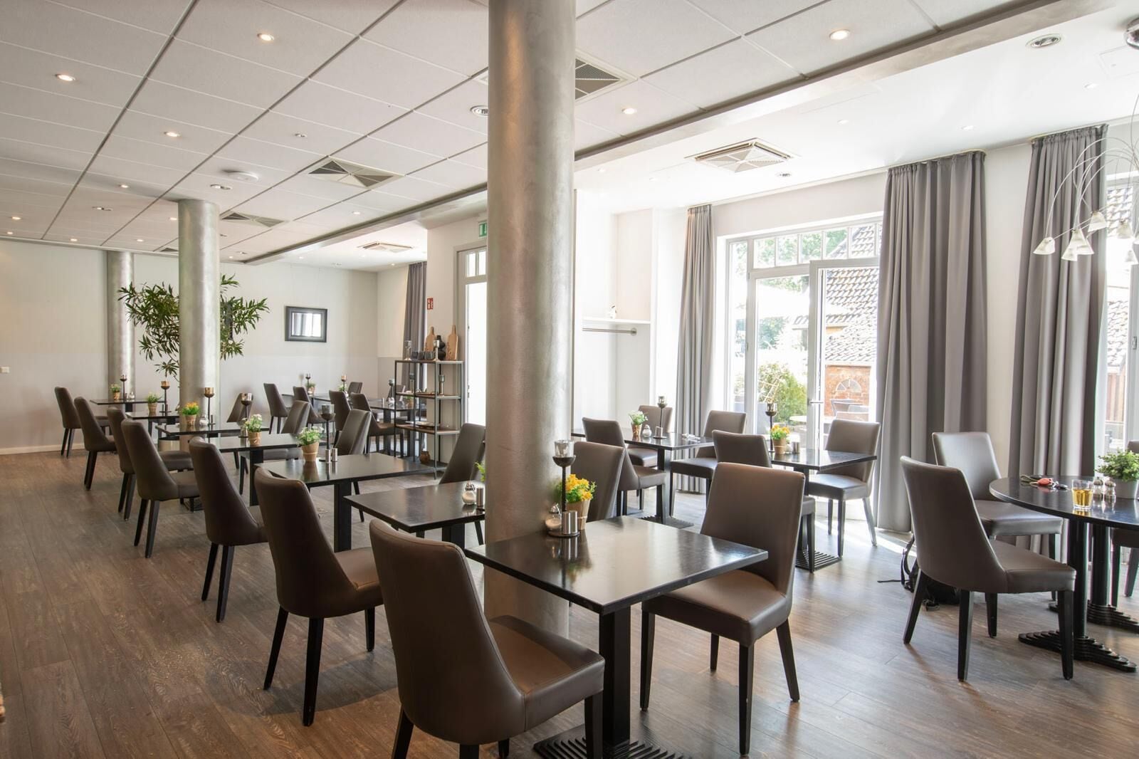 En rummelig spisesal med flere borde og stole, store vinduer med gardiner og indeplanter.