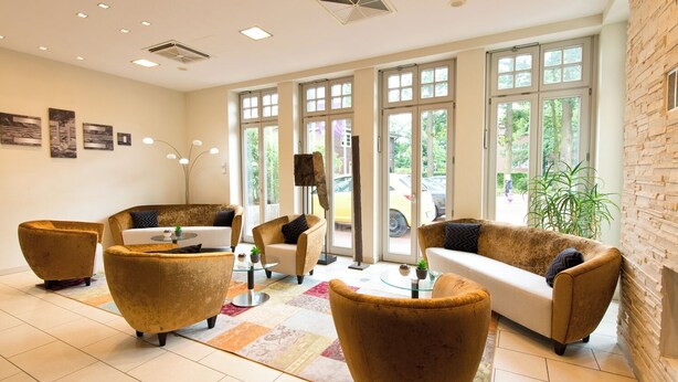 Un hall d’entrée spacieux, doté d’un design moderne, comprenant un canapé, des fauteuils et une table basse. De grandes fenêtres laissent entrer la lumière naturelle et offrent une vue sur l’extérieur.