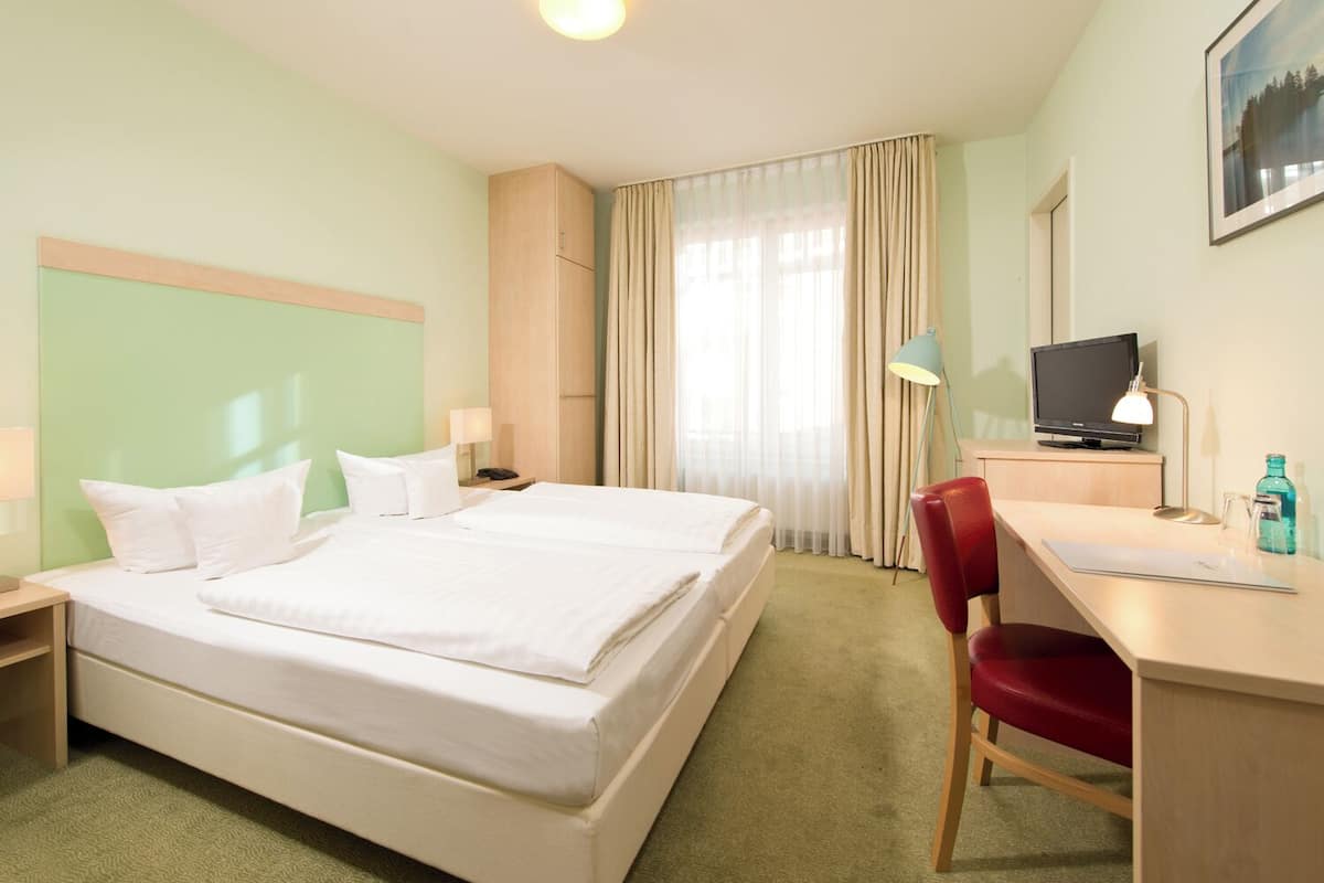 Habitación de hotel con cama, escritorio, silla, televisión, lámpara y cortinas.