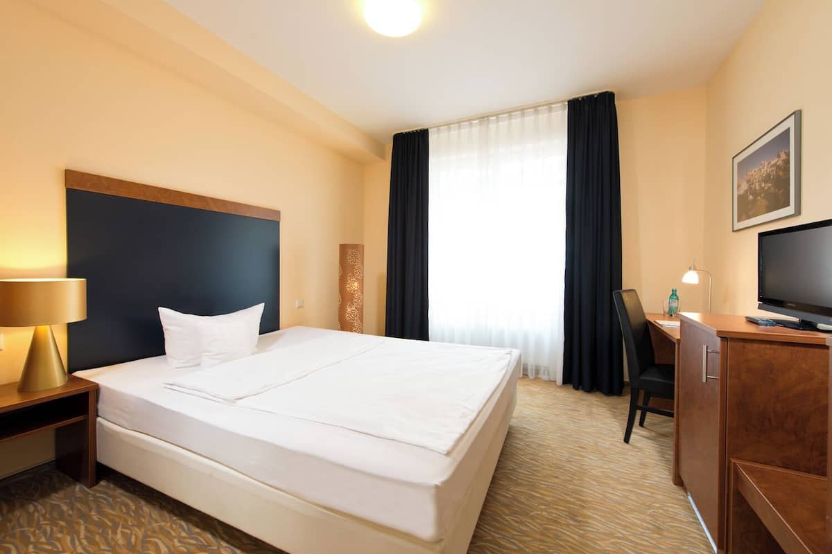 Una habitación de hotel con una cama grande, un escritorio con computadora, una lámpara y un televisor.