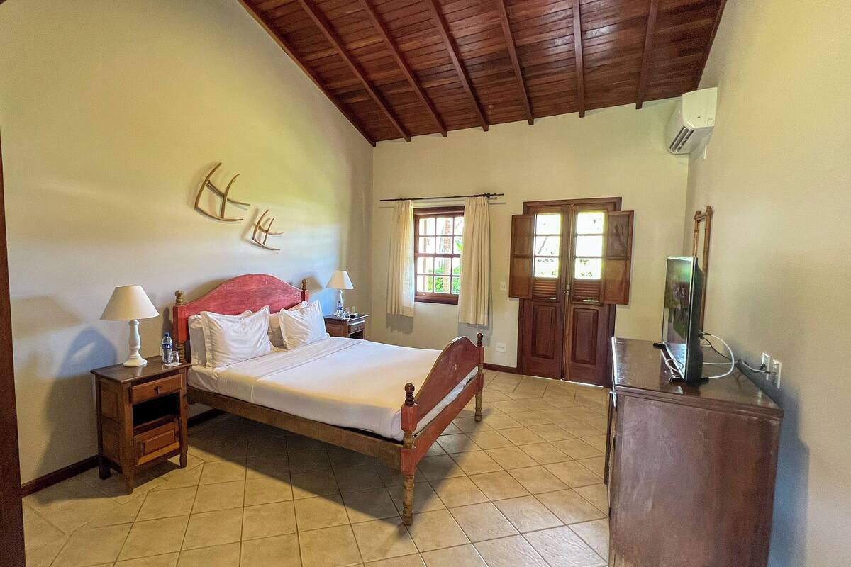 Un dormitorio con cama de madera, una mesita de noche, un mueble para televisor, una ventana con cortinas y techo de madera.