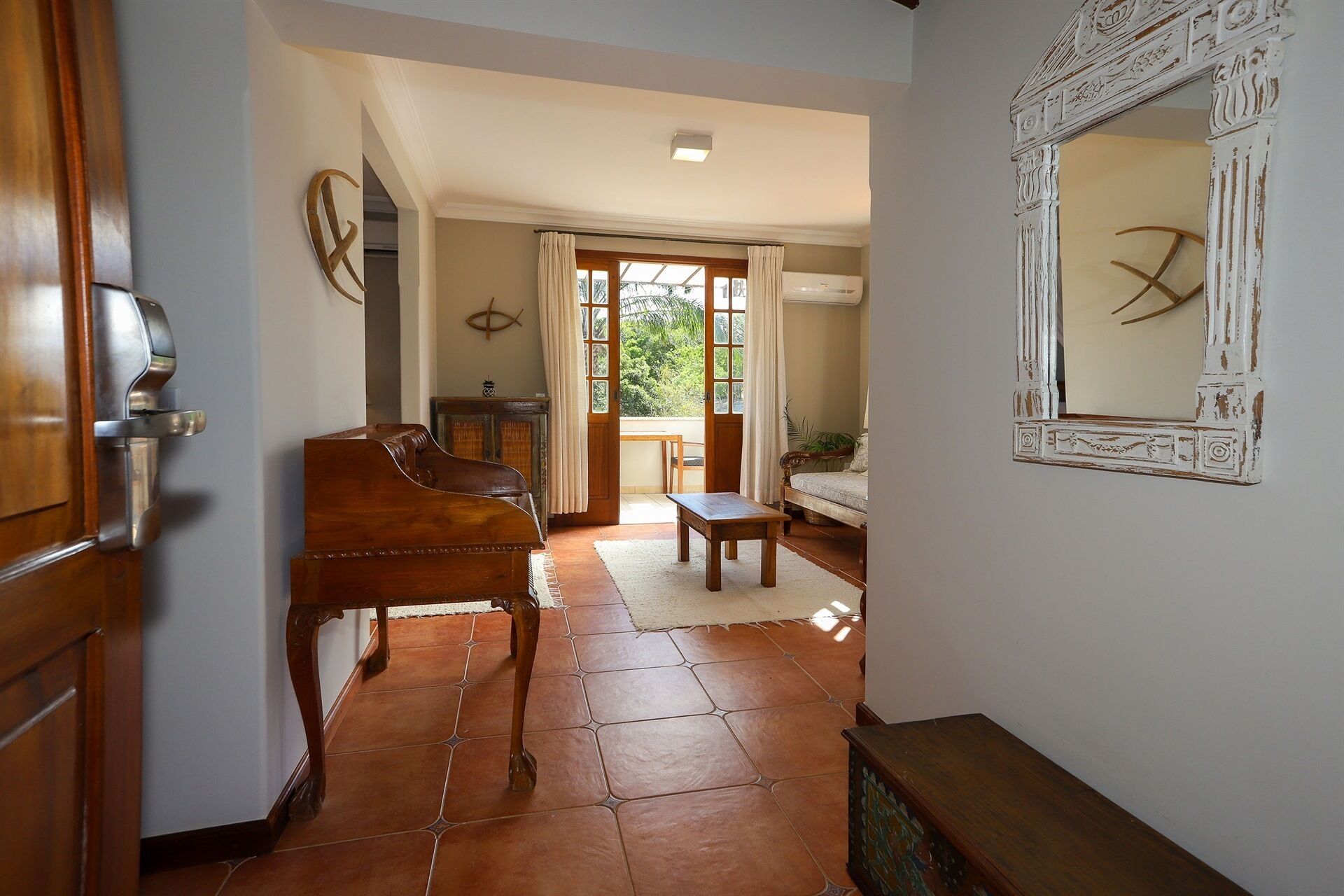 A hallway with a piano, a mirror, and a view into a room with a bed and curtains.
