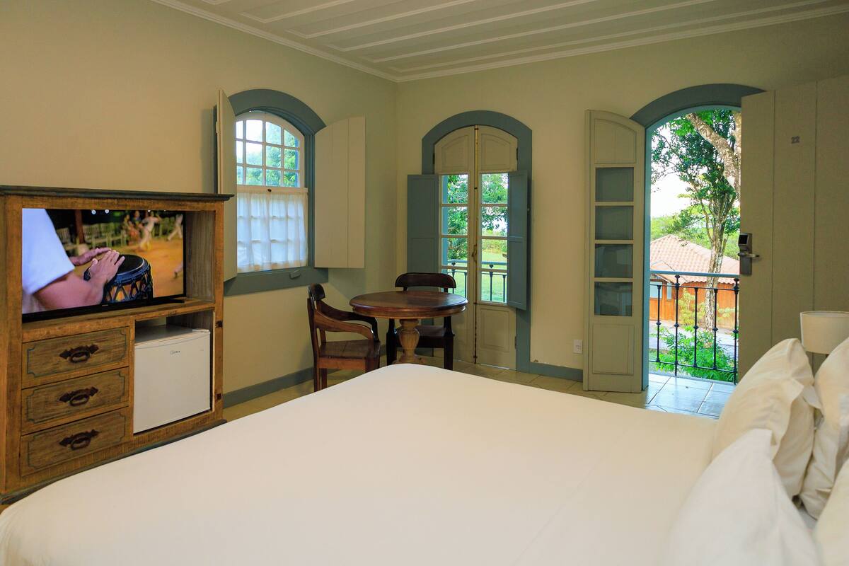 Un dormitorio con cama, televisor, una mesita y sillas. Hay dos ventanas con contraventanas, una de ellas con vista al exterior.