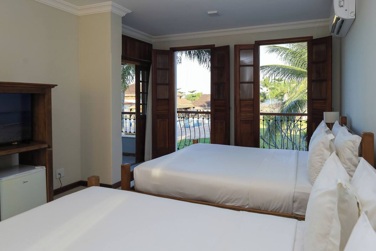 Habitación de hotel con dos camas, televisión y balcón con vistas a palmeras y una piscina.